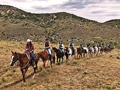 A wrangler leads a long group of riders along a dusty mountain trail.
