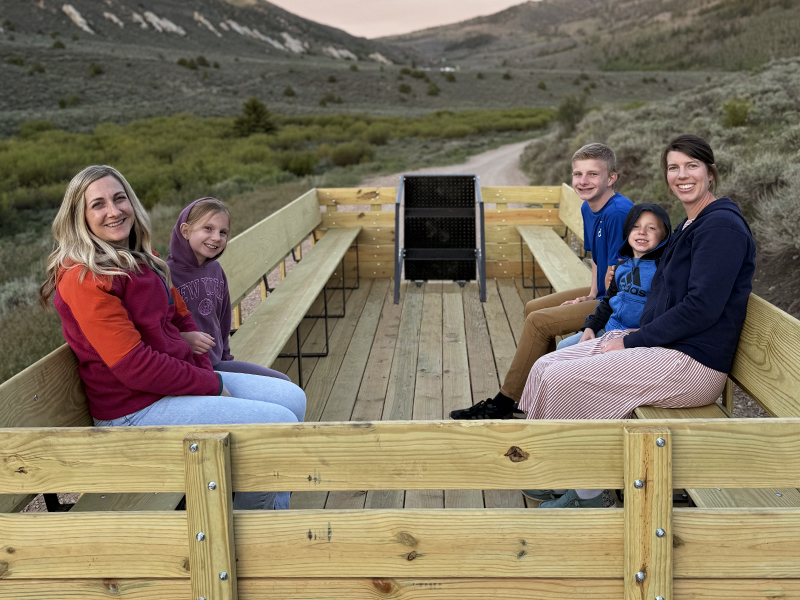Looking back at the wagon as it is pulled by a truck over the dirt road. Seated on wood benches guests enjoy the ride.